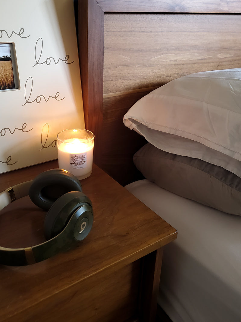 luxury crystal infused water sign candle on wood nightstand with beats headphones beside bed with grey and white linens