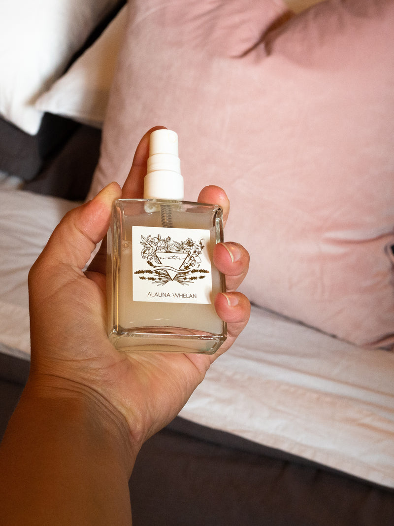 hand holding water sign aromatic ritual mist with grey and white bedding and pink velvet cushion in background