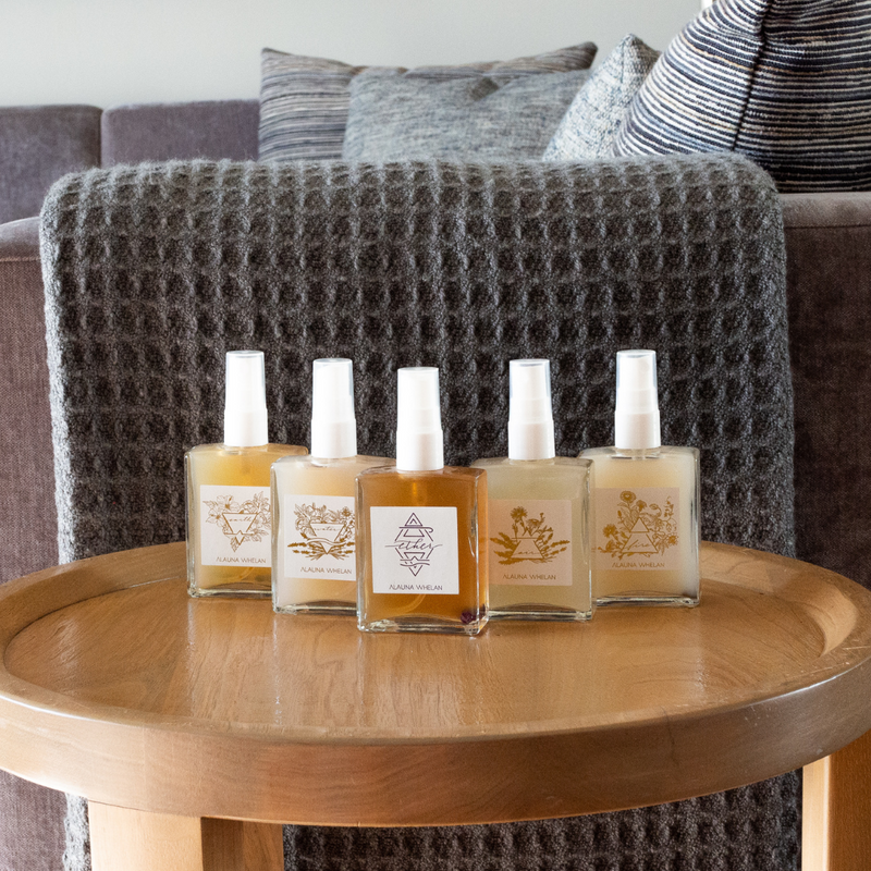 five glass ritual mist bottles on wooden table in front of a grey couch with pillows and a throw blanket
