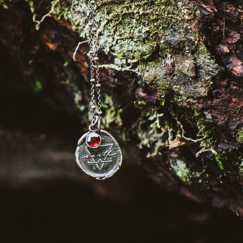 silver earth element medallion talisman necklace with red gemstone and moss and foliage in the background