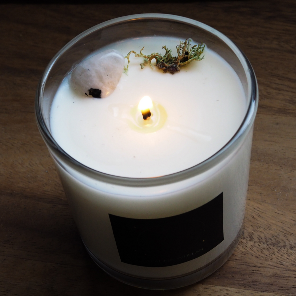 birds eye view of lit new moon soy candle topped with white moonstone crystal and moss on wooden tray